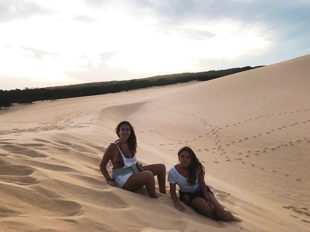 Dos chicas morenas senetadas en el desierto sonriendo a cámara